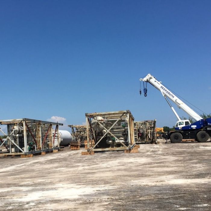 Large industrial equipment modules, likely for oil and gas processing, are stored in an expansive outdoor laydown yard under a clear blue sky, with a white and blue crane in the background.