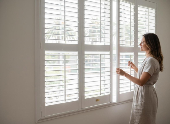 A woman in a linen dress opens white plantation shutters, revealing a view of green trees and palm leaves outside a bright window. Woman opening white window shutters
