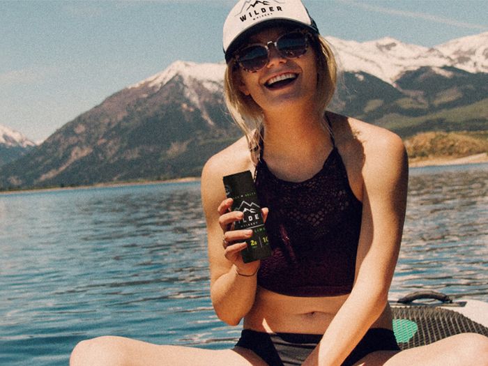 A young woman enjoys a Wilder Whiskey seltzer by a lake.