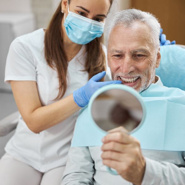 A male patient in a dental chair looks into a hand mirror, while a masked dental assistant in blue gloves adjusts his mouth. A male patient in a dental chair looks into a hand mirror, while a masked dental assistant in blue gloves adjusts his mouth.