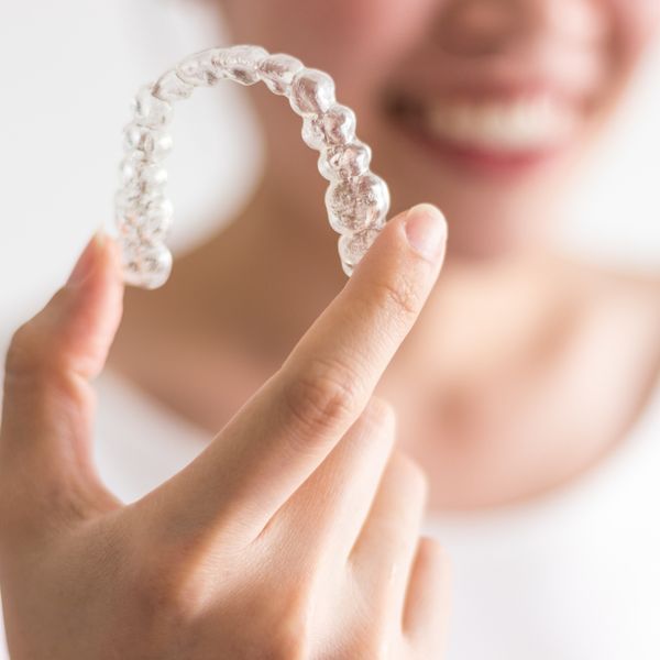 A person's hand holds a clear Invisalign aligner close-up, with a blurred smiling face in the background.