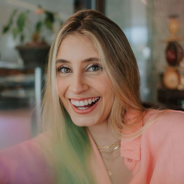 A young woman with long blonde hair and green eyes smiles broadly at the camera, wearing a pink shirt.