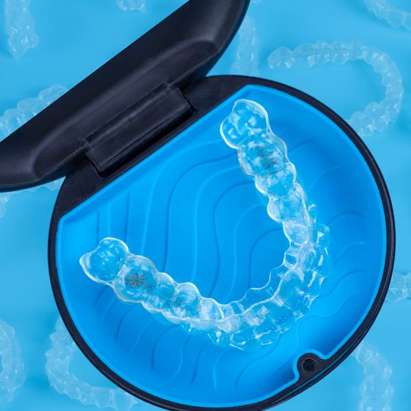 A clear Invisalign aligner rests inside an open black and blue carrying case, with other blurred aligners scattered on a blue background.