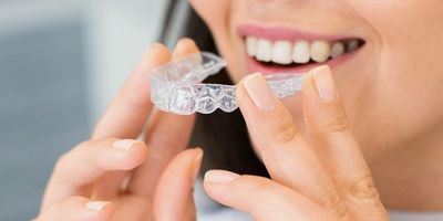 A close-up of a person smiling, holding a clear Invisalign aligner near their mouth, showing white teeth.