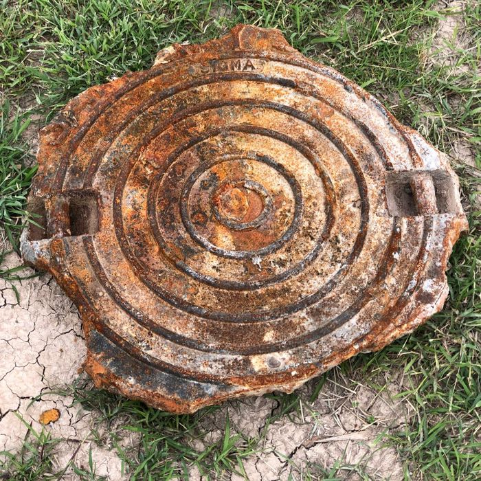 A single, severely rusted and weathered iron manhole cover lying on dry, cracked ground. A single, severely rusted and weathered iron manhole cover lying on dry, cracked ground.