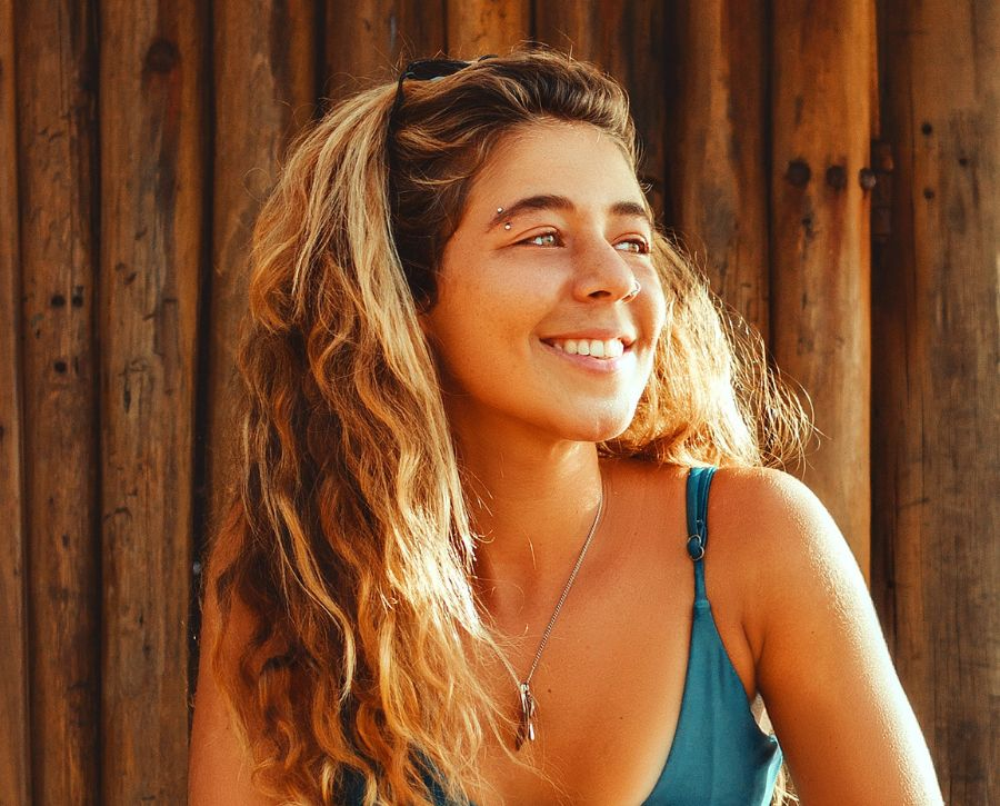 A young woman with long, wavy blonde hair and a healthy smile stands in the sun against a wooden wall.