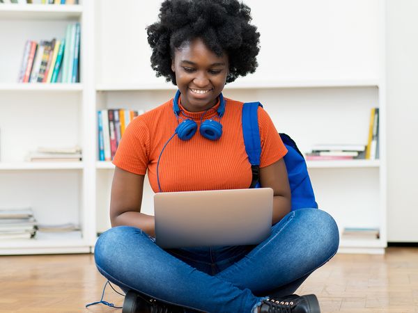 African American girl sitting on the floor on a laptop with headphones around her neck, smiling. 