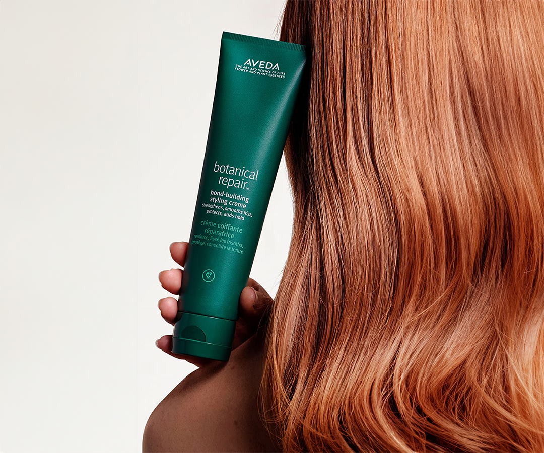 Picture of woman with red hair holding bottle of Aveda's botanical repair creme Hair Treatments Image