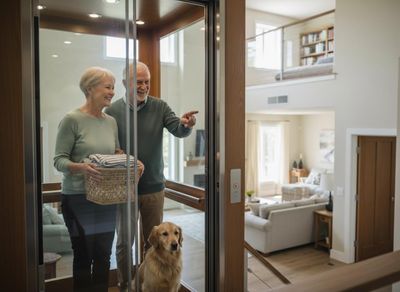 A happy senior couple and their golden retriever ride a glass home elevator, looking out into their bright, modern house. Senior Couple and Dog in Home Elevator