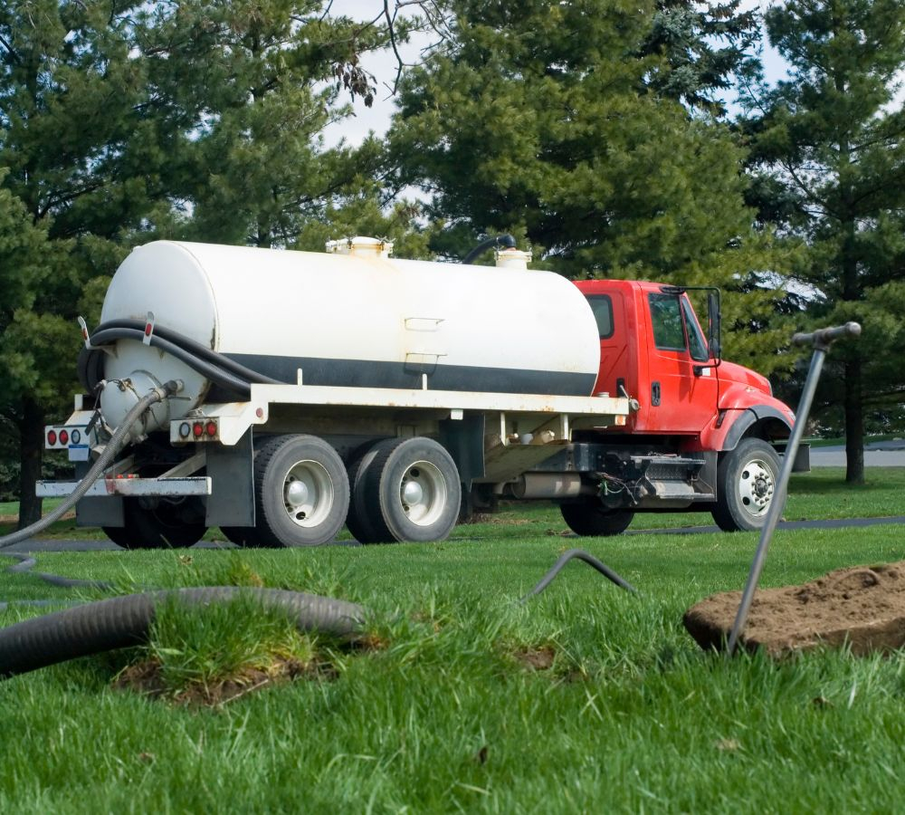 a septic truck in front of a hold in the ground with a hose running into the hole