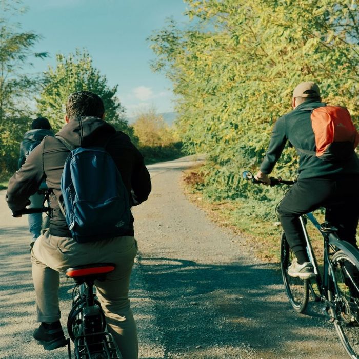 Three people with backpacks ride their bicycles down a sunlit, paved nature trail surrounded by trees.