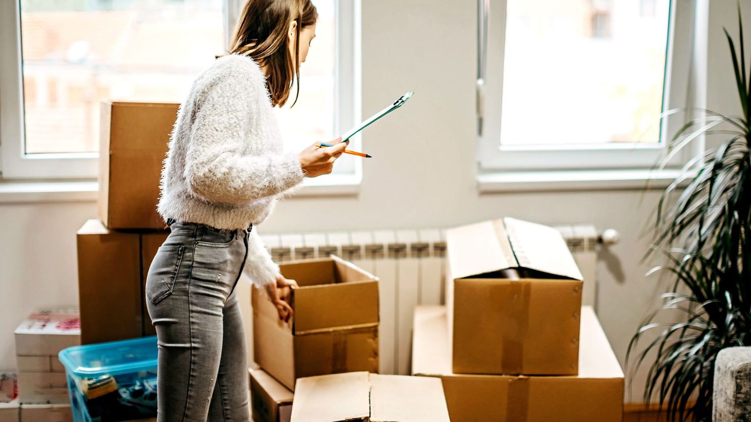 Person holding a clipboard and looking at moving boxes stacking in their apartment