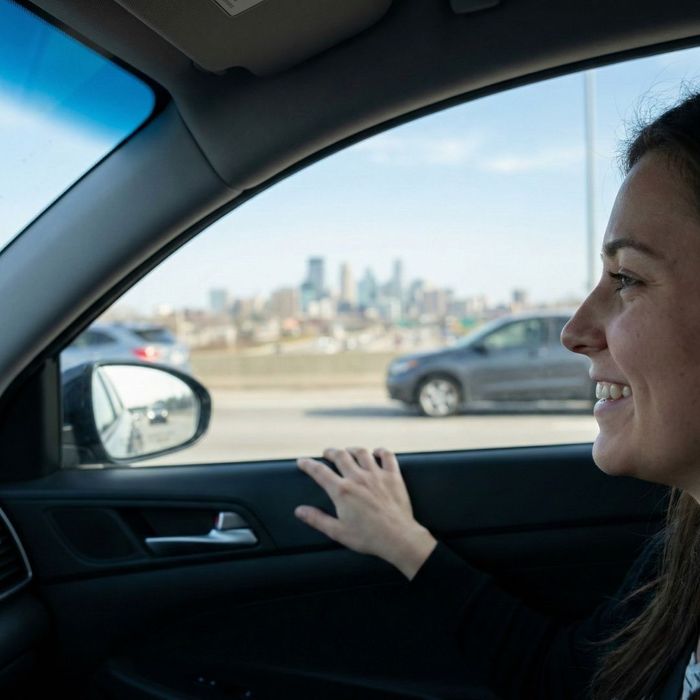 A woman smiles while looking out of a car window at a blurred city skyline in the distance during a daytime drive.