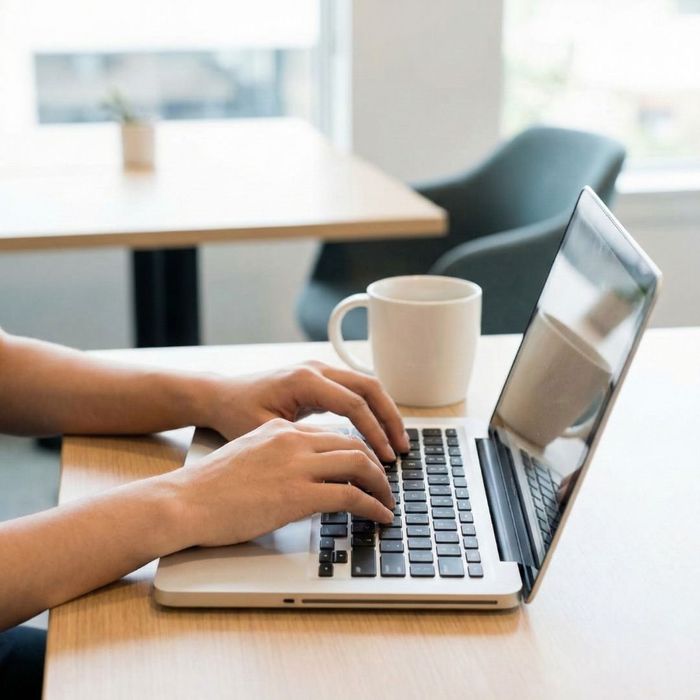 A close-up view of hands typing on a silver laptop at a wooden desk in a bright, modern indoor workspace with a white coffee mug nearby.