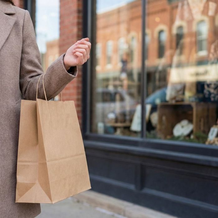 A close-up view of a person in a tan coat holding a brown paper shopping bag while standing in front of a historic brick storefront window.