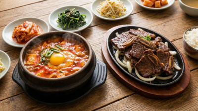 spread of authentic Korean dishes including a steaming pot of Sundubu and a plate of Galbi ribs on a wooden table