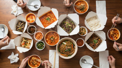 photograph captures a wooden table filled with various open white takeout containers holding Korean dishes like kimchi stew, bulgogi, and noodles, with several hands reaching in with chopsticks