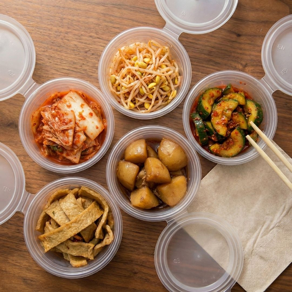 five small, round plastic containers arranged on a wooden table, each holding a different Korean side dish