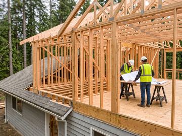 Two contractors in safety vests standing on a new second-story home subfloor and reviewing architectural blueprints.