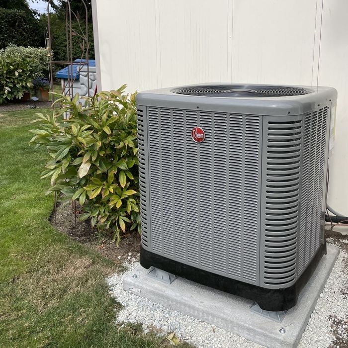 A gray outdoor air conditioning unit with a Rheem logo sits on a concrete pad surrounded by gravel and grass, next to a leafy green bush.