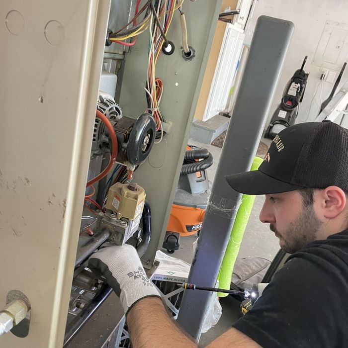 A professional HVAC technician wearing a black cap and work gloves carefully inspecting and repairing the internal wiring and motor of an open furnace unit.