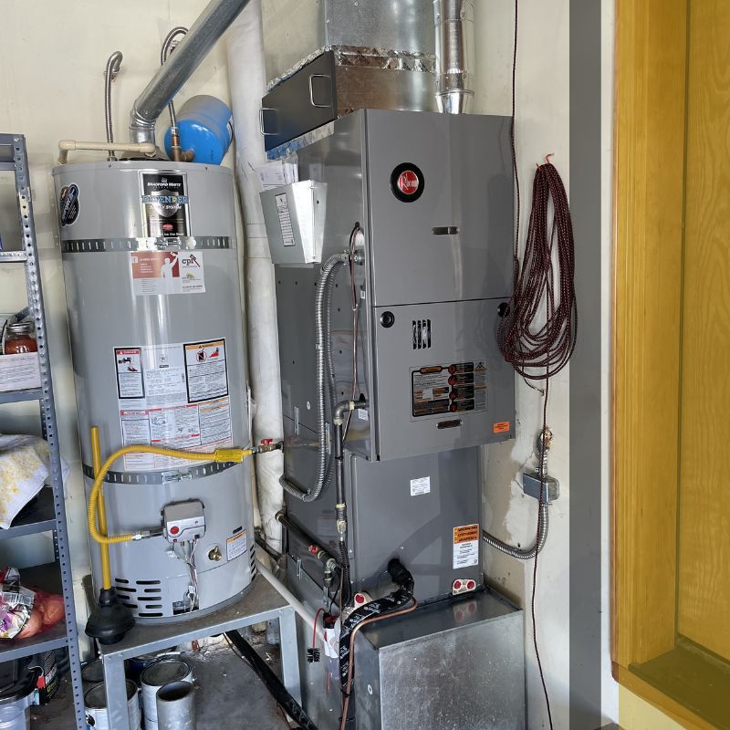 A gray gas furnace and a gray water heater are installed side-by-side, connected by various pipes and ducts.