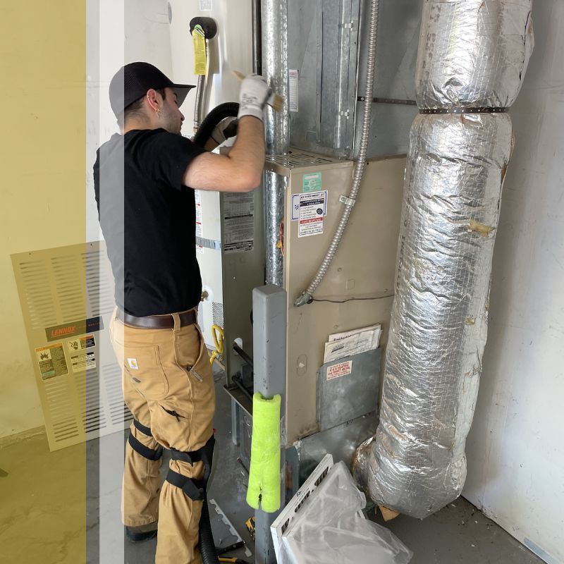 A person wearing a black shirt and tan work pants is working on an HVAC unit. A large air filter is leaning against the wall to the left of the unit.
