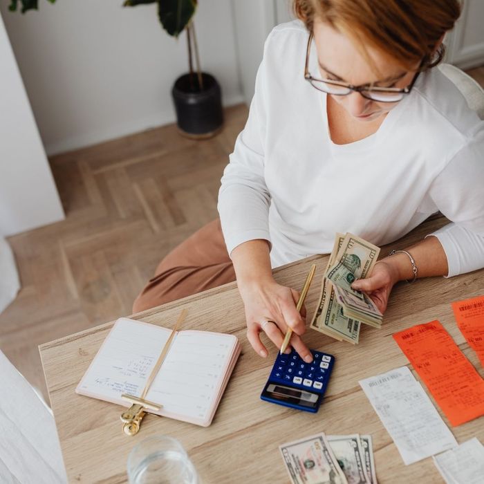 A top-down view of a woman using a calculator and counting cash next to a pile of red receipts and an open notebook on a wooden desk.