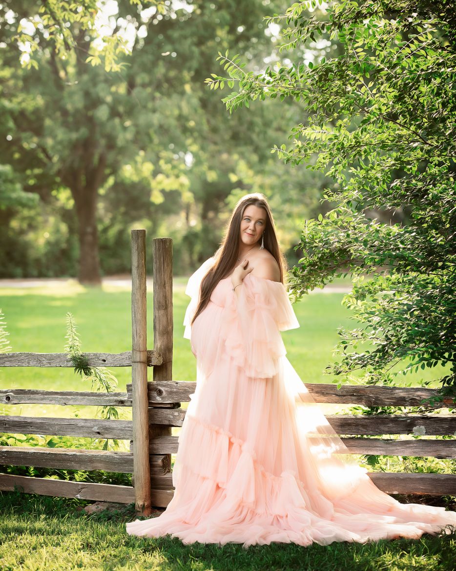 Expectant mother in flowing blush gown beside rustic fence outdoors