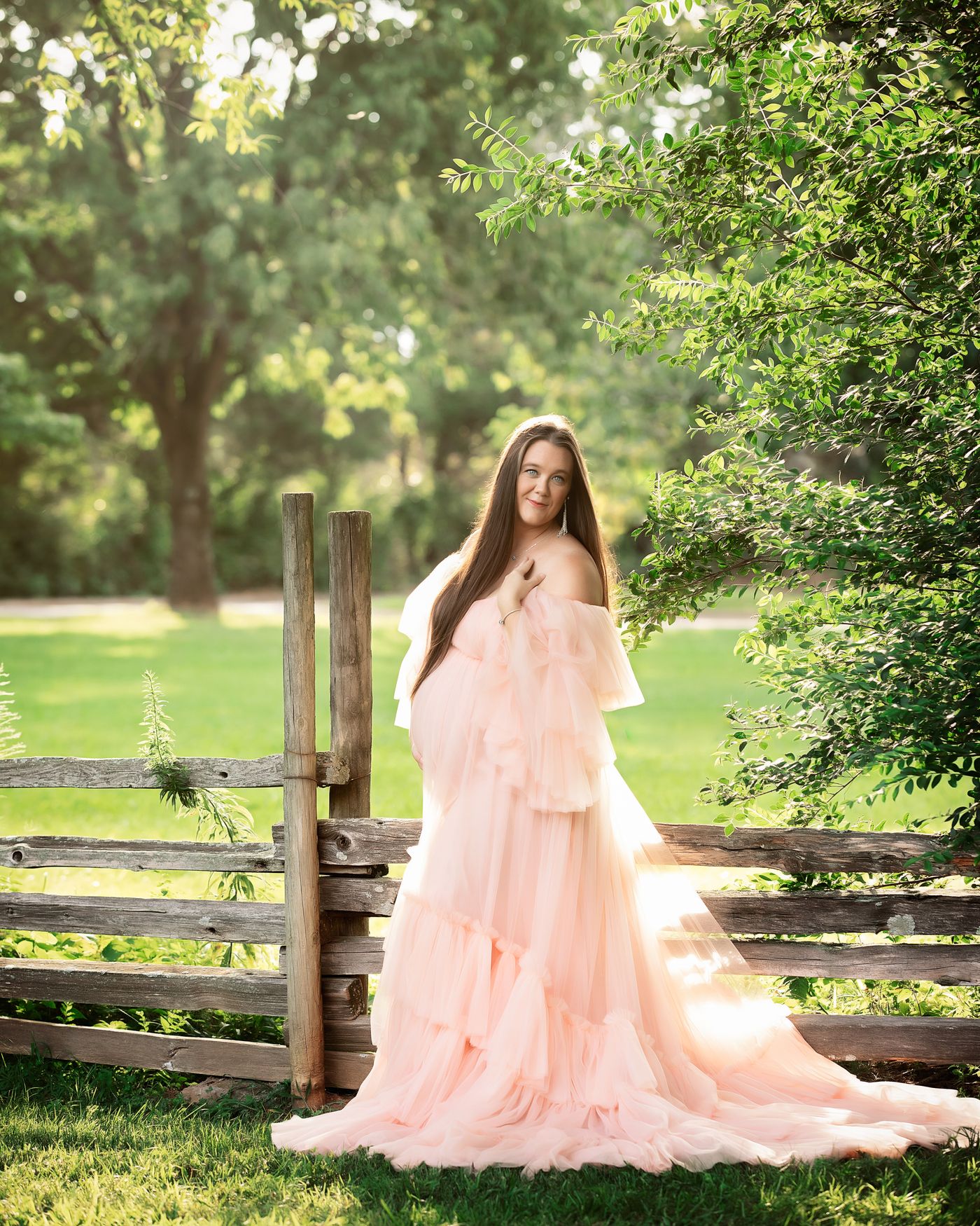 Expectant mother in flowing blush gown beside rustic fence outdoors