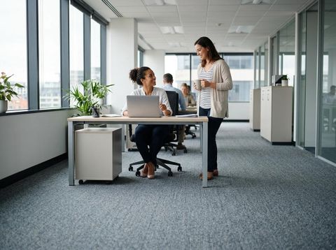 Two women, one seated at a laptop, one standing with a mug, smiling and talking in a bright, open-plan office with large windows. Colleagues collaborating in a modern office
