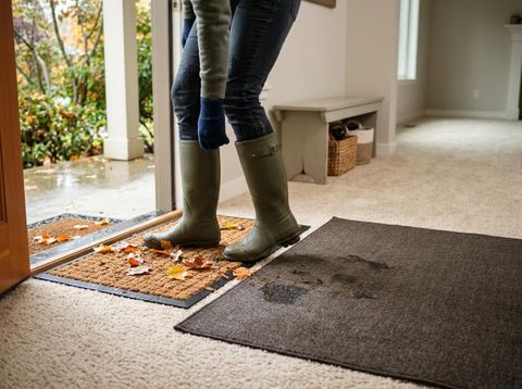 A person in green rain boots and jeans steps from a wet outdoors onto a coir doormat with leaves. Muddy footprints mark a mat inside. Person stepping inside with muddy boots on a doormat