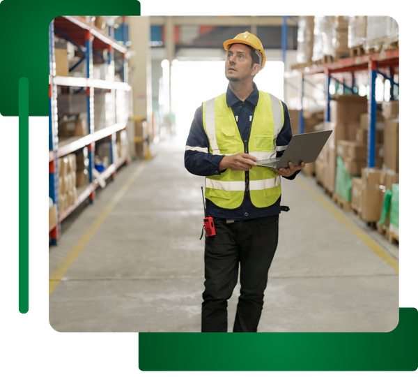 Man in safety vest/helmet standing in a warehouse aisle holding a laptop, looking up.