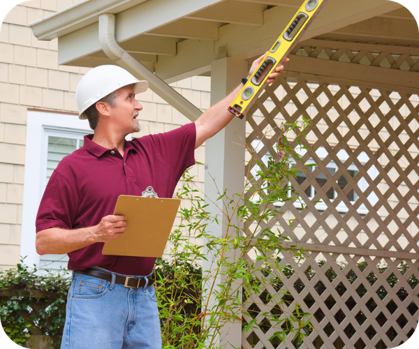 Home inspector in a hard hat holding a level tool and clipboard, inspecting the exterior of a house.