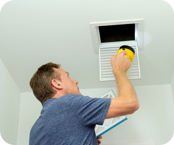 Home inspector using a flashlight to examine an air vent and filter in a ceiling.