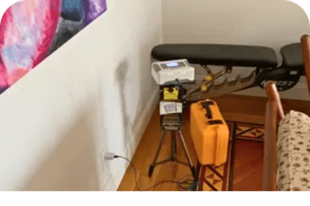 Radon testing equipment set up on a tripod in a room corner, next to a wall outlet.