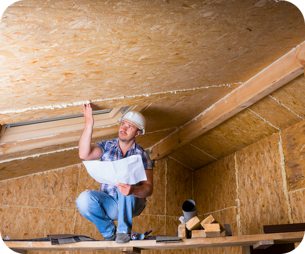 Construction worker in a hard hat crouching on scaffolding, inspecting a ceiling or roof structure with blueprints.