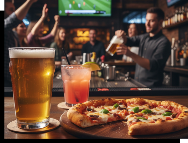A close-up of a pizza slice, a beer, and a cocktail on a bar counter, with a blurred background showing people watching a soccer game on TV and a bartender serving drinks.
