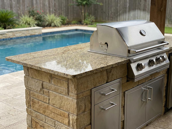 Close-up of a custom Houston outdoor kitchen with a granite countertop, stacked-stone base, and stainless steel built-in grill beside a pool