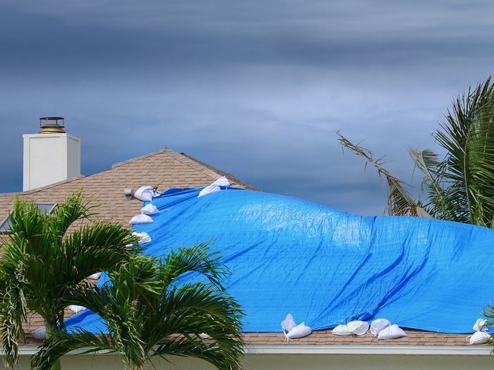  Storm damaged roof on house with a blue plastic tarp over hole in the rooftop.