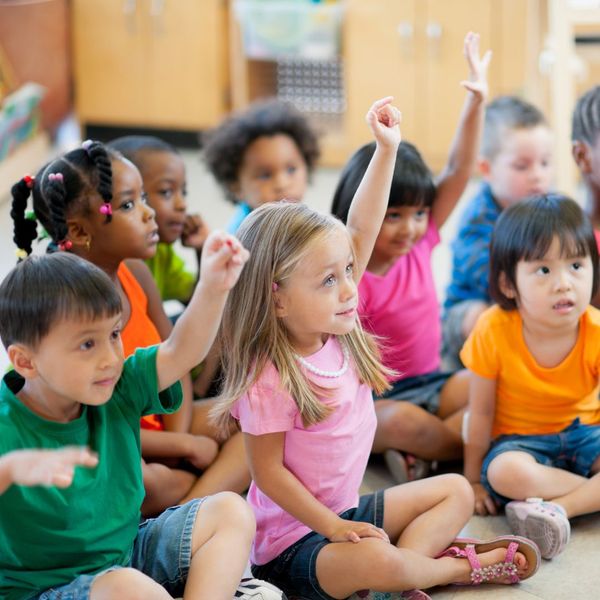 classroom of preschool students sitting on the floor and raising their hands