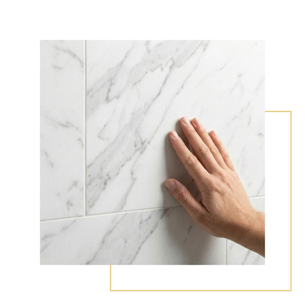 A hand gently touching the smooth surface of a high-quality white marble shower tile with grey veining.