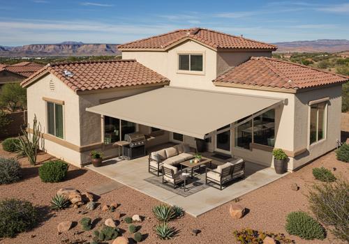 High-angle aerial view of a desert modern home with a large tan retractable fabric awning extended over a concrete patio layout with outdoor seating and a grill. An elegant home with a retractable patio awning and an outdoor living area in Southern UtahHigh-angle aerial view of a desert modern home with a large tan retractable fabric awning extended over a concrete patio layout with outdoor seating and a grill.