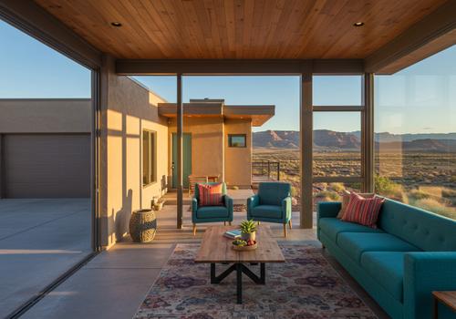 Interior of a luxury home featuring a retractable glass wall opening onto a patio. The room has mid-century modern furniture, a wood-slat ceiling, and expansive windows showcasing the red rock mountains of Southern Utah. Luxury Home Living Area with Desert View