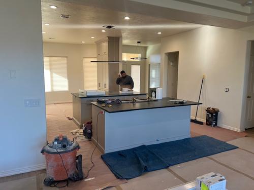 Modern kitchen remodel under construction featuring dual islands with dark countertops, recessed ceiling lights, protective floor coverings, and a worker onsite finalizing details. Kitchen Countertop Professional Install
