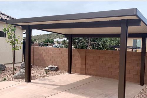 Modern solid top aluminum patio cover and carport structure featuring dark brown steel support posts and trim, installed over a concrete pad in a residential backyard with a block retaining wall and desert landscaping. A newly installed patio cover in st george, utah with a carport design, featuring a metal roof and brown columns, with a tiled wall in the background.