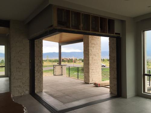 This new home construction project showcases a stunning transition from indoor to outdoor living with a large bifolding glass door. The design includes beautiful stone columns and a natural wood ceiling on the covered patio, perfectly framing the scenic view of the St. George mountains. This type of feature is ideal for creating an open, airy living space. New home construction with large bifold doors, a stone accent wall, and a view of the mountains.