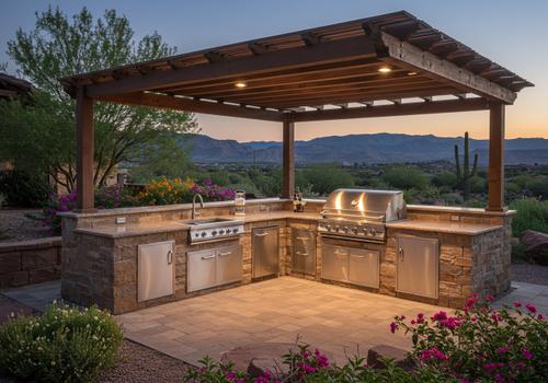 Custom outdoor kitchen and L-shaped stone barbecue island built under a dark stained wood pergola with integrated lighting, featuring a premium stainless steel grill, sink, under-counter storage doors, and a scenic desert mountain background at dusk. outdoor kitchen design