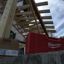 Low-angle close-up view of an outdoor patio pergola wooden framing installation project under a cloudy sky, with a red Milwaukee tool box visible in the foreground. framing contractor
