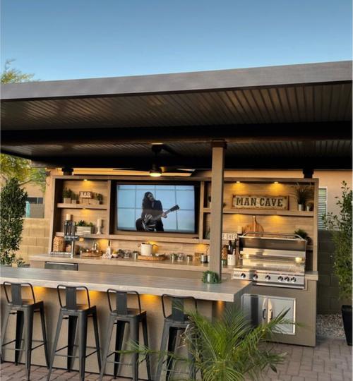 High-end outdoor entertainment kitchen and bar island under a solid patio cover, featuring wood panel shelving with accent lighting, a large flat-screen TV, built-in stainless steel grill, and black metal bar stools. modern outdoor entertainment area